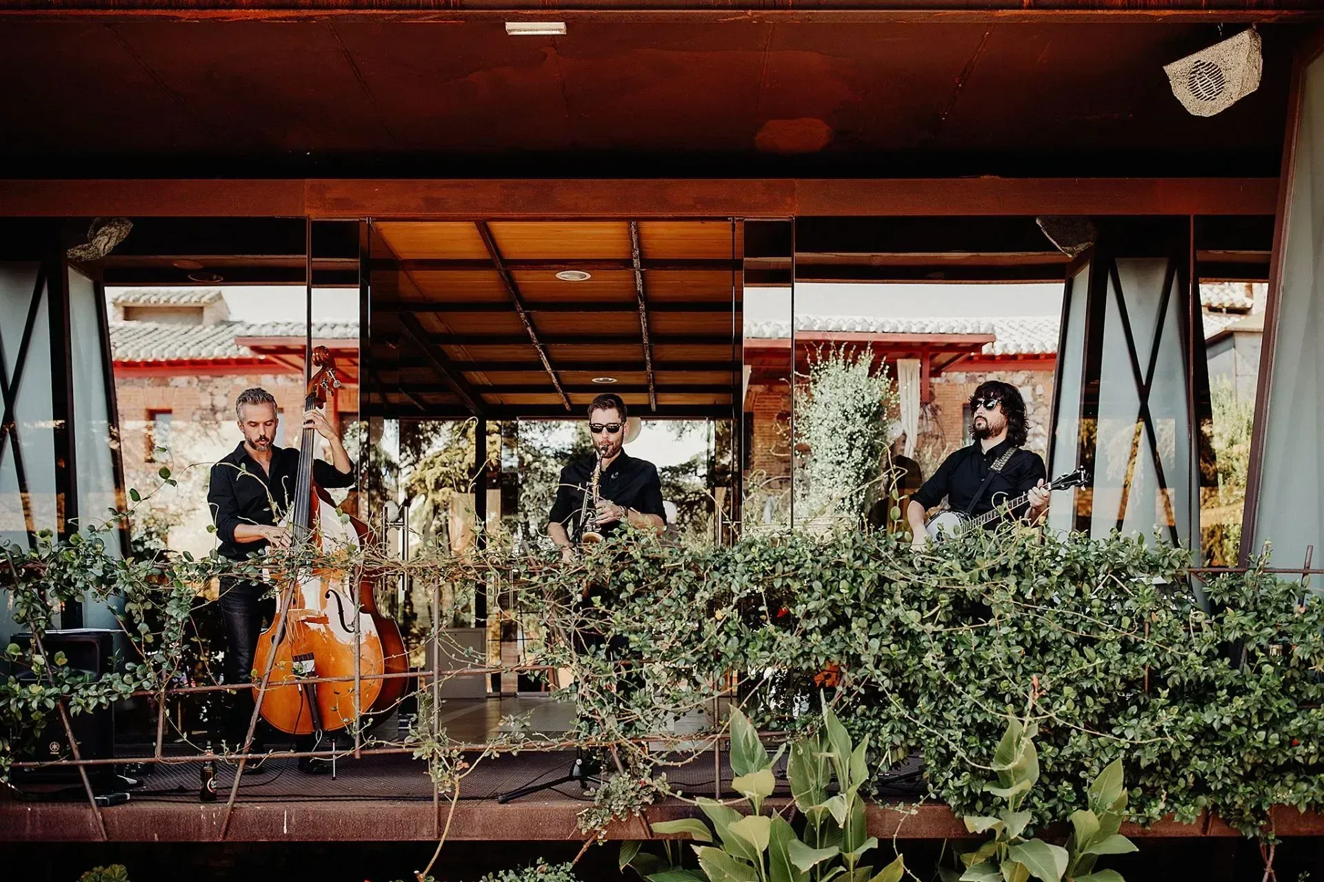 Banda tocando en un balcón: tres hombres con instrumentos. Al aire libre, rodeados de vegetación.