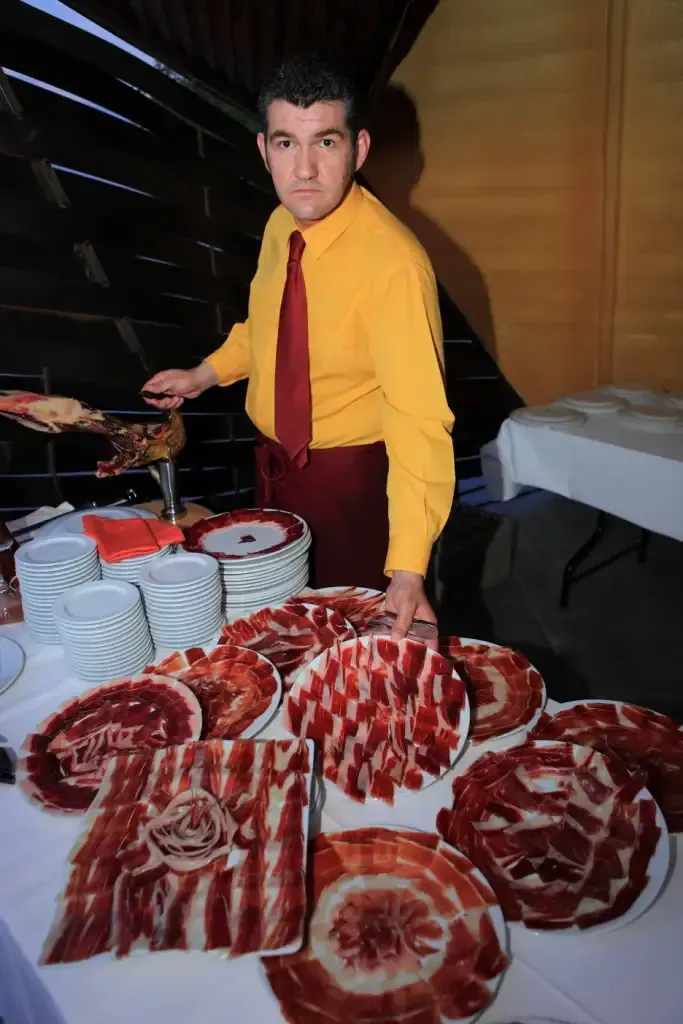 Hombre con camisa amarilla cortando jamón en una mesa con platos de jamón en lonchas.