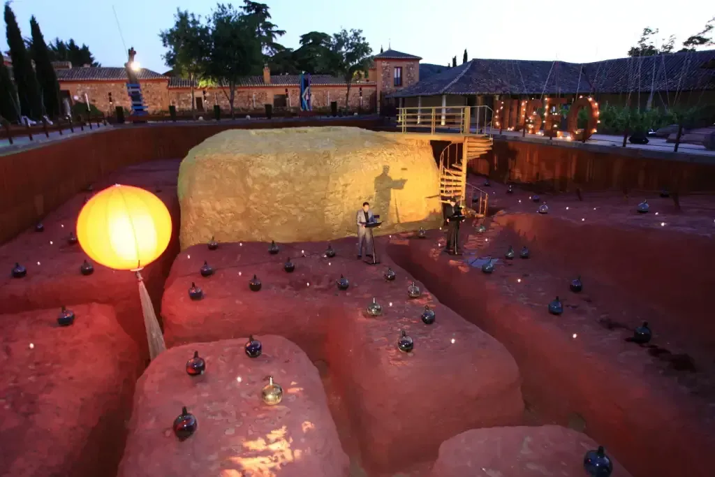 Instalación de arte al aire libre al anochecer, con montículos de tierra, luces, un fardo de heno y una estructura; una linterna brilla.