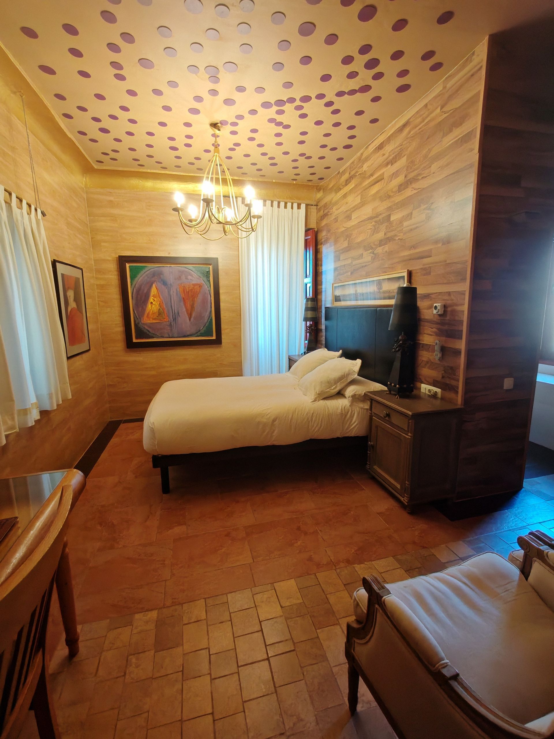 Dormitorio con paredes revestidas de madera, ropa de cama blanca y una lámpara de araña; puertas francesas que se abren a la luz del sol.