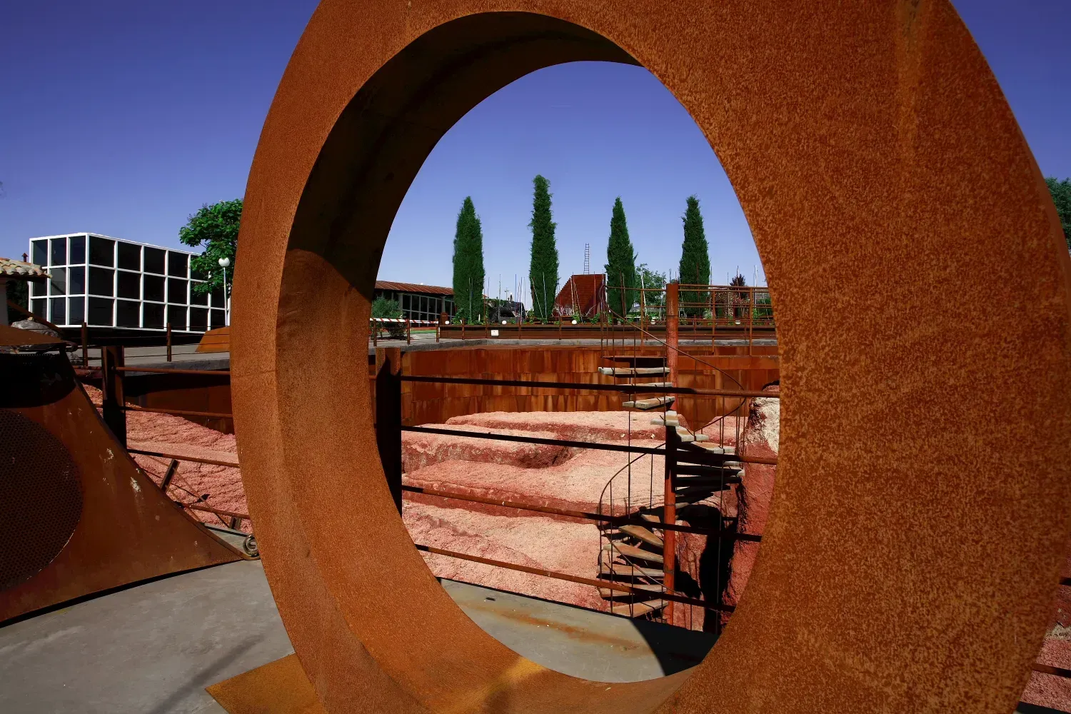 Una gran escultura de metal oxidado enmarca una vista de edificios y árboles altos contra un cielo azul.