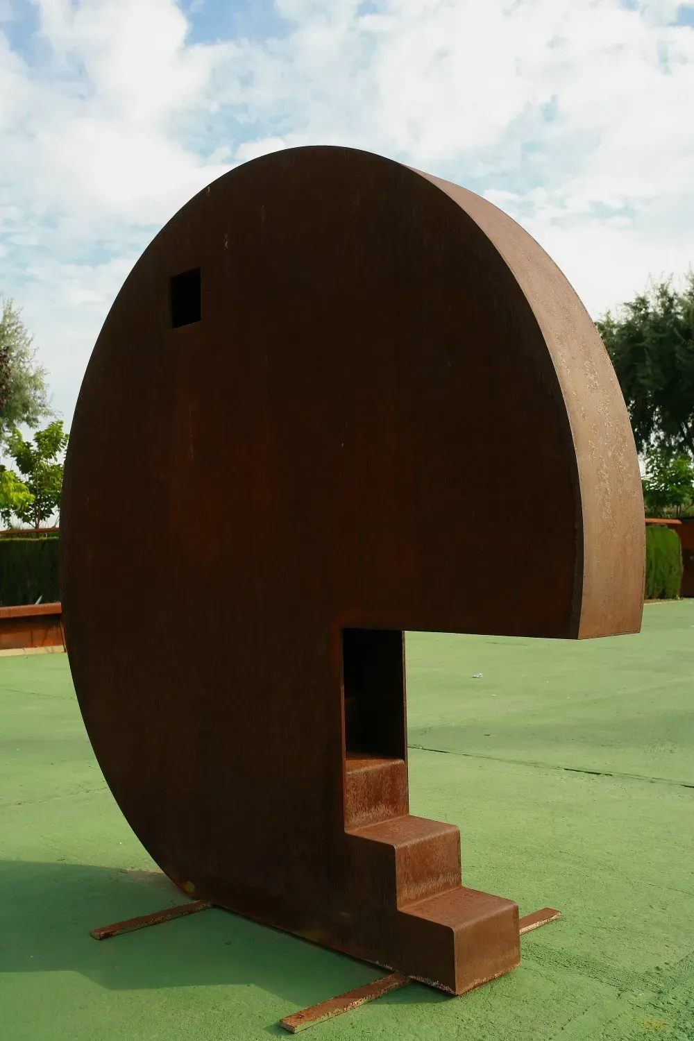 Gran escultura circular de acero de color marrón con una sección recortada y escalones sobre suelo verde.