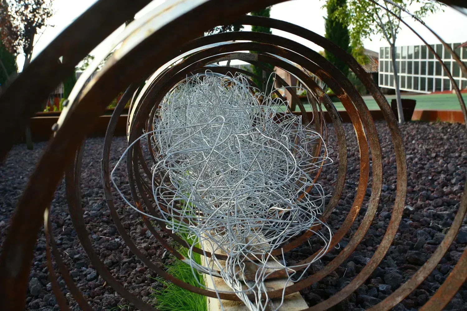 Anillos de metal que se enrollan hacia adentro y enmarcan una masa enredada de cables plateados, en un jardín con rocas marrones.