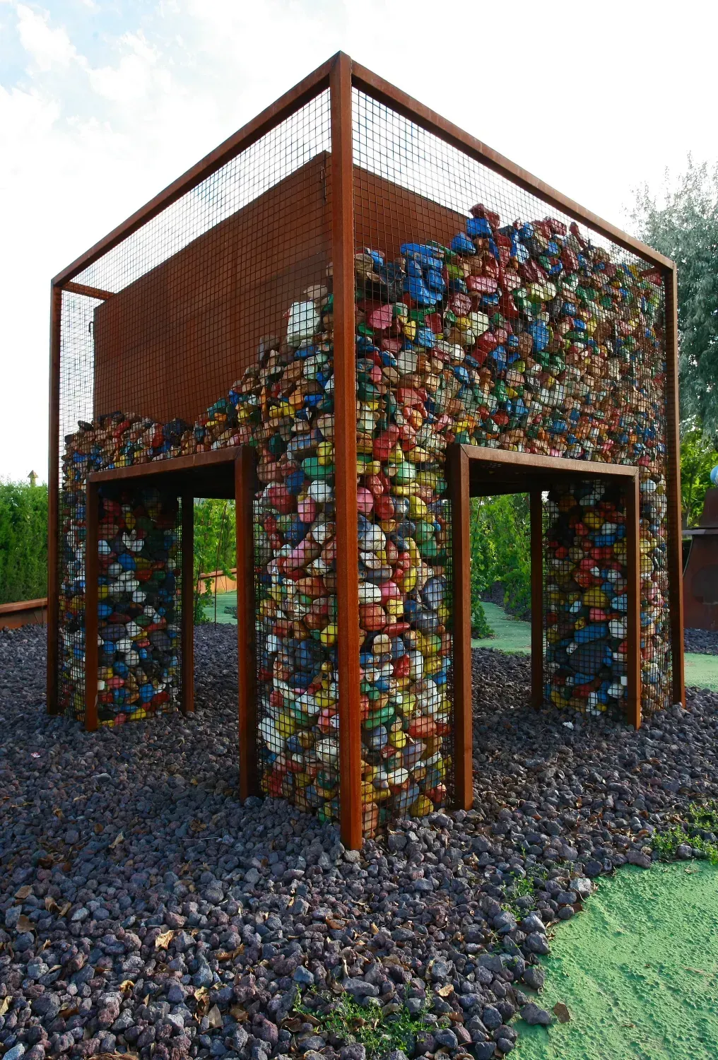 Escultura al aire libre: Estructura de jaula de metal llena de objetos coloridos y redondeados, colocada sobre grava oscura.