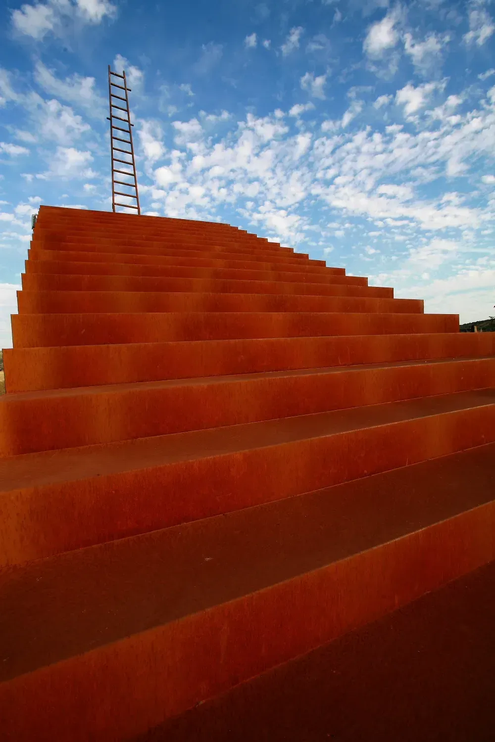 Gran escalera naranja que conduce al cielo, con una escalera de metal encima contra un cielo azul y nublado.