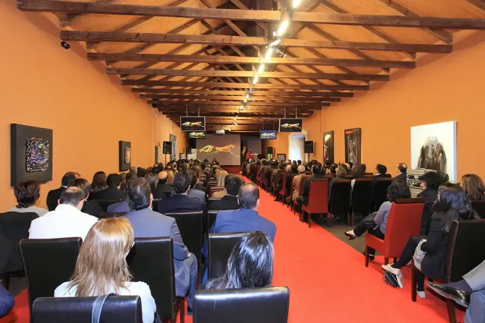 Público sentado en un largo salón para una presentación, alfombra roja, paredes naranjas, techo de madera y pantallas.