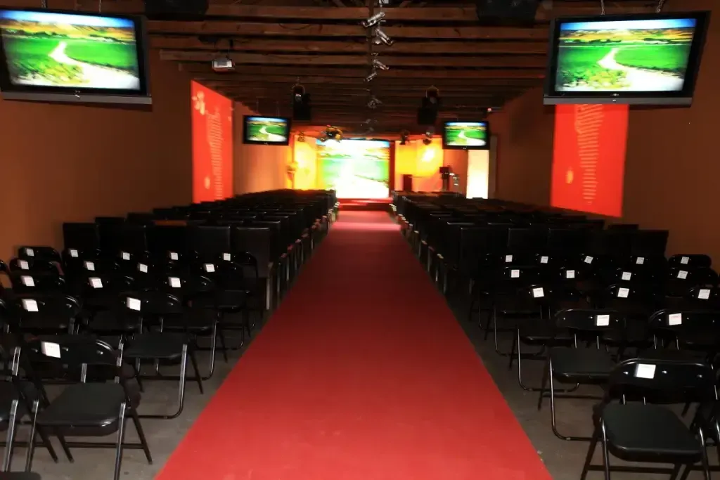 Pasarela vacía con alfombra roja, flanqueada por filas de sillas negras. Dos pantallas muestran el paisaje.