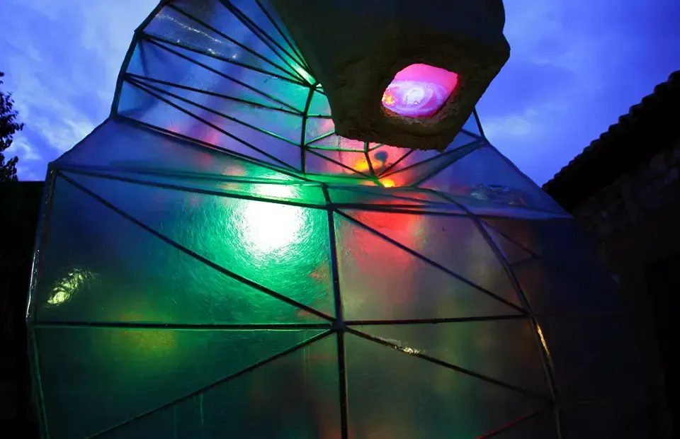 Gran escultura geométrica iluminada con luces internas de colores contra un cielo crepuscular.