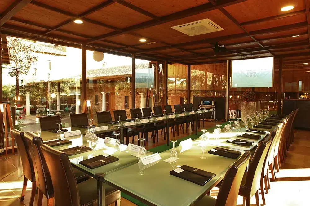 Sala de conferencias con mesa larga, sillas, computadoras portátiles y pantalla; ventanas grandes; techo de madera.