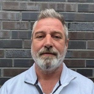 Ein Mann mit Bart steht vor einer Backsteinmauer.