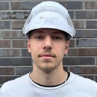 Ein junger Mann mit Schutzhelm und Schutzbrille steht vor einer Backsteinmauer.