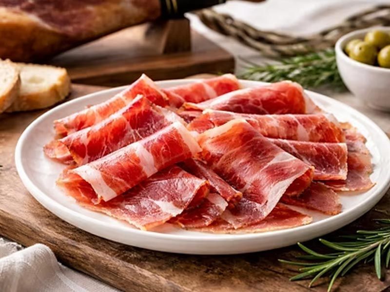 Plato de jamón curado en lonchas finas sobre una tabla de madera con romero y aceitunas al lado.