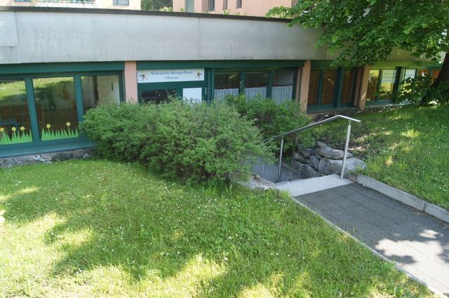 Ein Gebäude, zu dem eine Treppe hinaufführt, ist von Gras und Büschen umgeben.