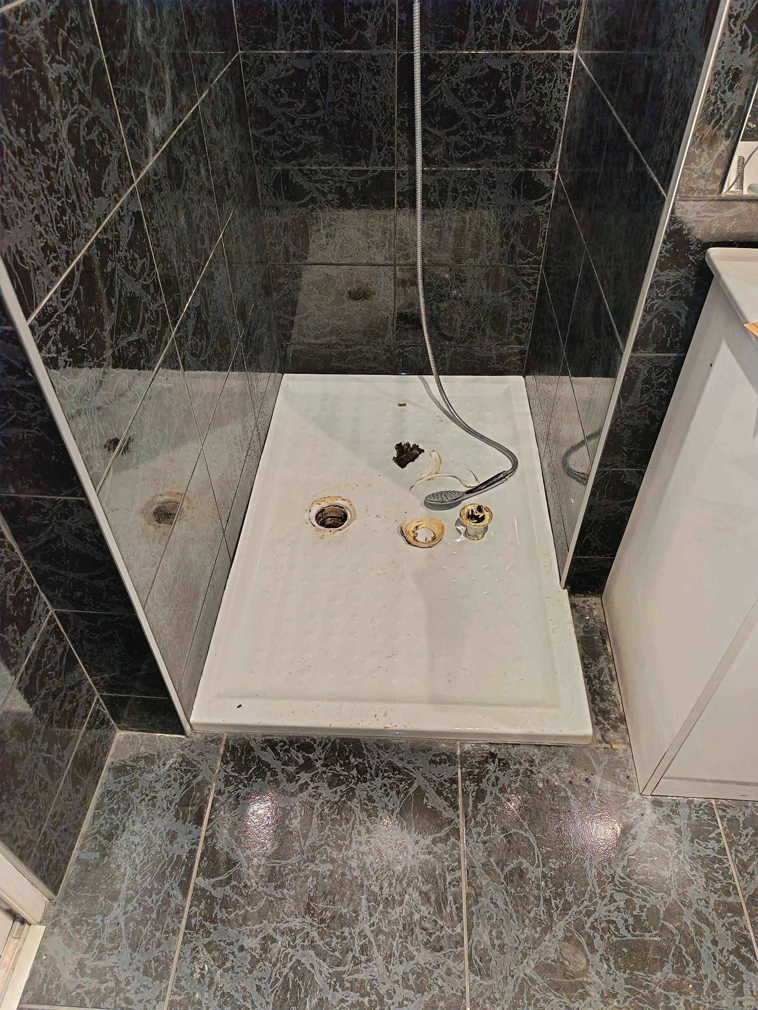 Douche avec murs et sol carrelés noirs ; receveur de douche blanc avec robinetterie.