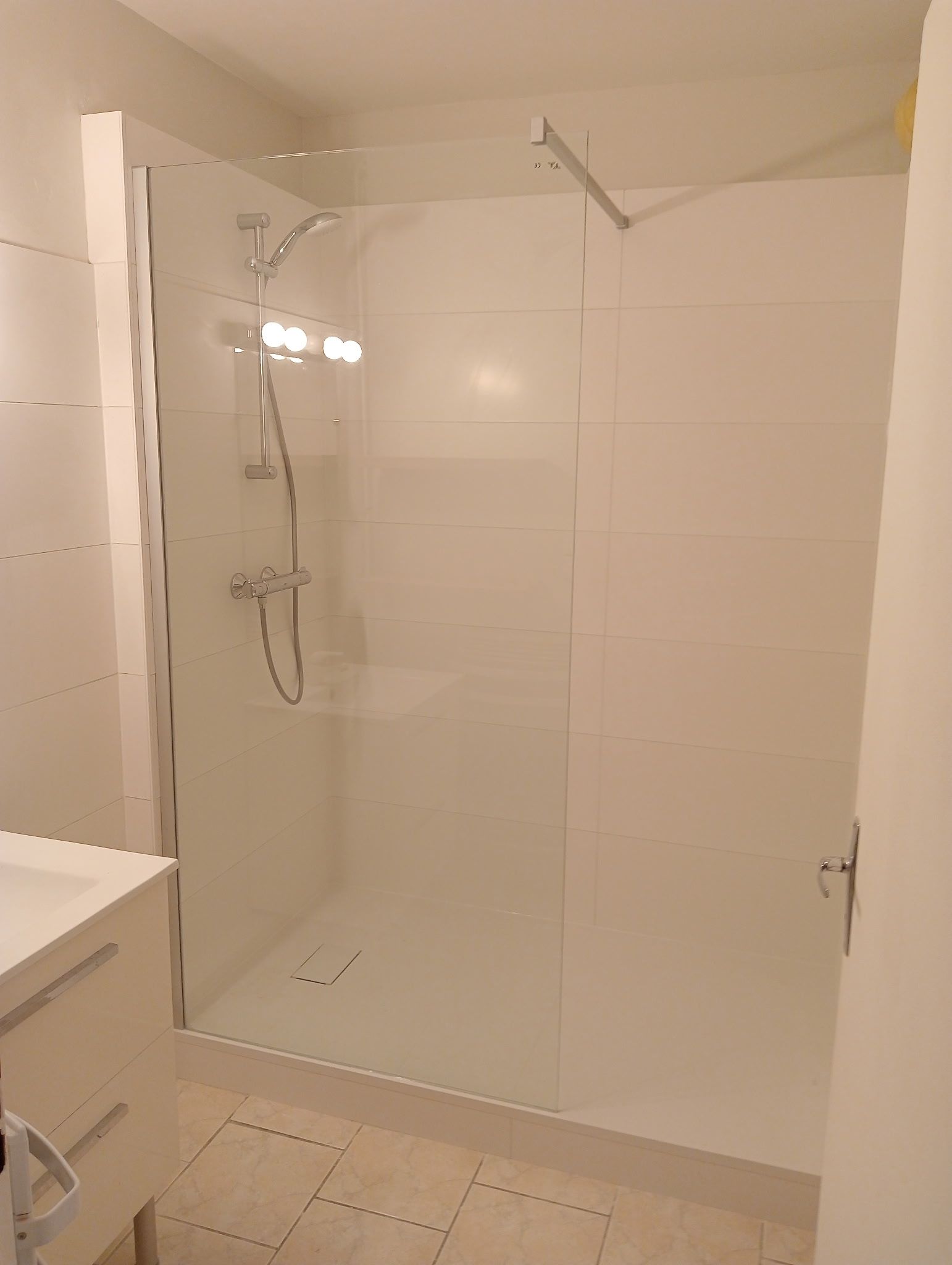 Cabine de douche avec murs carrelés blancs et porte en verre. Robinetterie chromée.