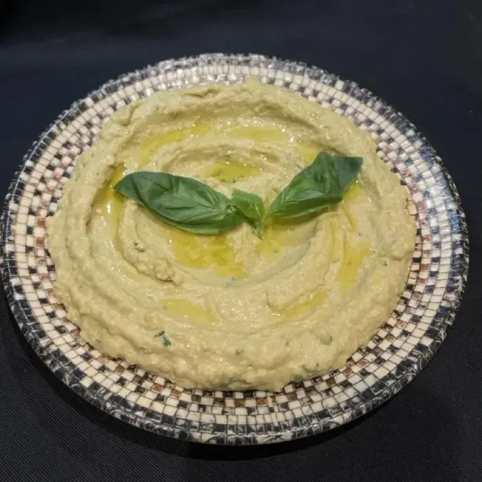 Eine Schüssel cremiger Hummus, garniert mit Olivenöl und Basilikumblättern, auf einem dekorativen Teller.