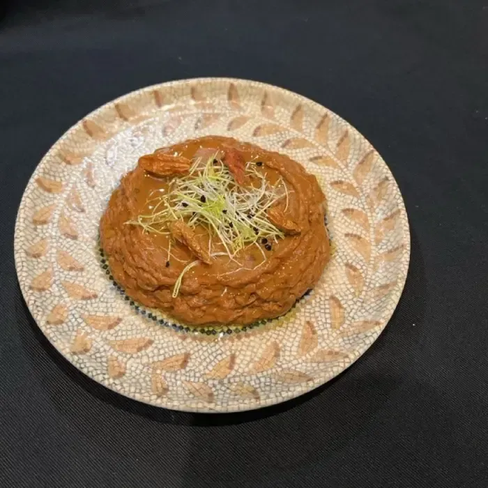 Ein auf einem gemusterten Teller vor dunklem Hintergrund angerichtetes, rot-oranges Currygericht, garniert mit Sprossen.
