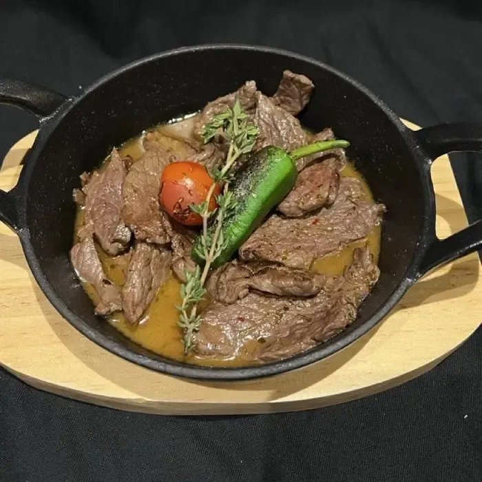 Schwarze Pfanne mit Rindfleischeintopf, grüner Paprika, Tomaten und Kräutern auf einem Holzbrett