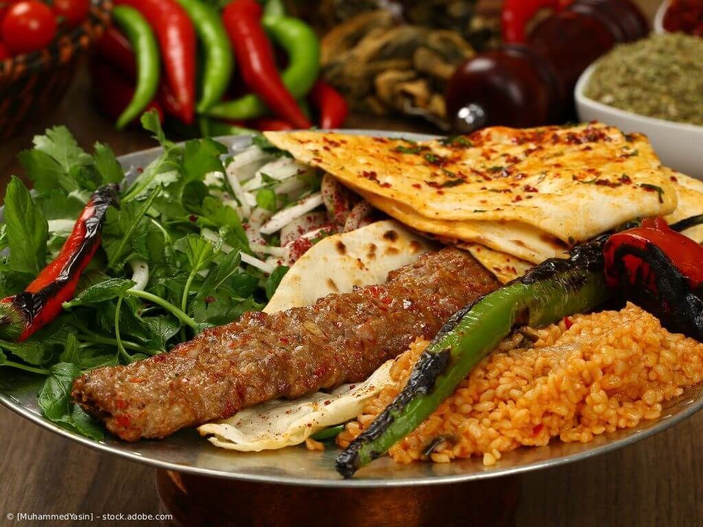 Gegrilltes Fleisch und Fladenbrot auf einem Teller mit Gemüse, Tomaten und Bulgur, dazu bunte Paprika.