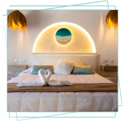 Una cama con dos cisnes hechos de toallas dispuestos en forma de corazón, con un cabecero semicircular retroiluminado con un diseño artístico de temática playera.
