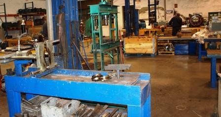 En el suelo de un taller industrial se observa un banco de trabajo azul en primer plano y una gran prensa de máquina verde en el centro.