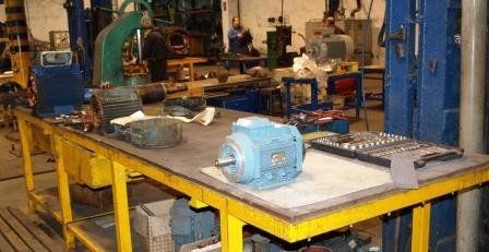 Un motor industrial azul descansa sobre un banco de trabajo metálico amarillo en un taller de maquinaria desordenado.