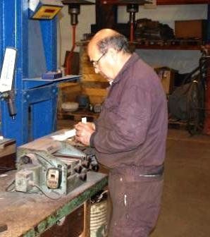 Una persona vestida con un mono marrón trabaja en una pequeña máquina en un taller, concentrada intensamente en un pequeño objeto que tiene en la mano.