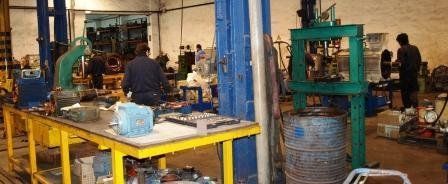 Los trabajadores manejan maquinaria en un taller industrial desordenado, con bancos de trabajo amarillos y una gran prensa verde.