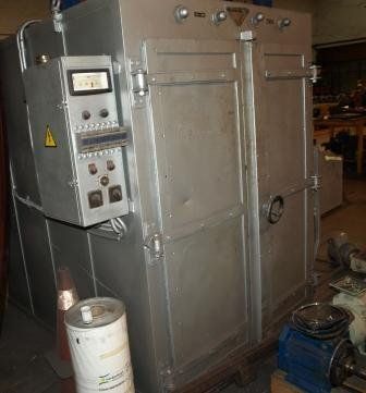 Un horno industrial metálico con panel de control frontal, situado en un taller con un tambor de almacenamiento y un motor cerca.