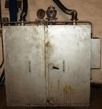 Un armario de secado de metal industrial, desgastado por el tiempo, con dos puertas frontales y conductos de ventilación, en un espacio utilitario con poca luz.