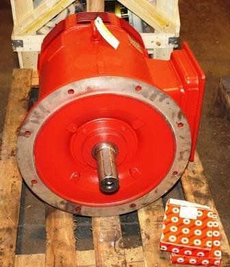 Un motor eléctrico industrial de color rojo brillante con un eje de salida con chaveta descansa sobre un palé de madera junto a un pequeño objeto embalado en una caja.