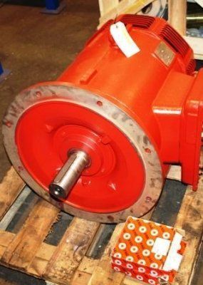 Un motor eléctrico industrial de color rojo brillante, con un eje de transmisión prominente, descansa sobre un palé de madera junto a un pequeño objeto embalado en una caja.