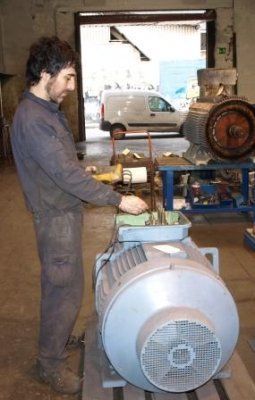 Un técnico con ropa de trabajo inspecciona la caja de bornes de un gran motor eléctrico industrial dentro de un taller de reparación.