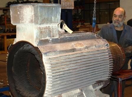 En un taller, una persona inspecciona la carcasa acanalada de un motor eléctrico industrial de gran tamaño que cuelga de un polipasto de cadena.