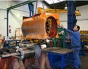 Un operario de taller guía un gran componente de una máquina industrial de color naranja, suspendido mediante un polipasto de cadena.