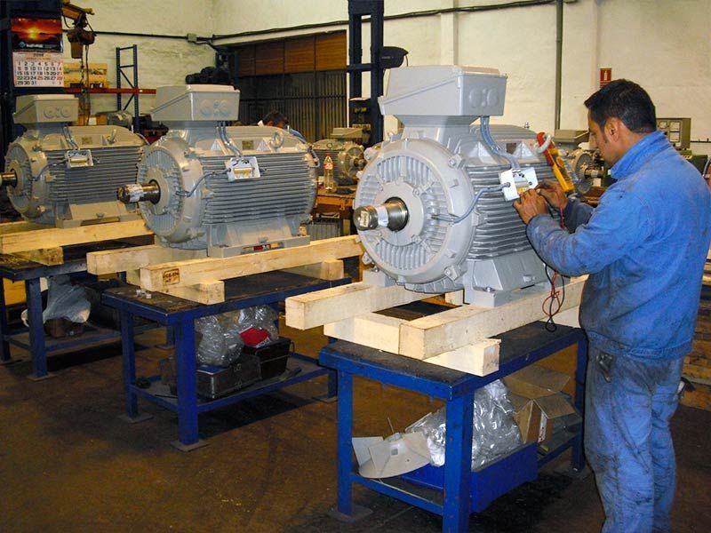Un técnico con uniforme azul utiliza un multímetro para probar un gran motor industrial gris en un taller.