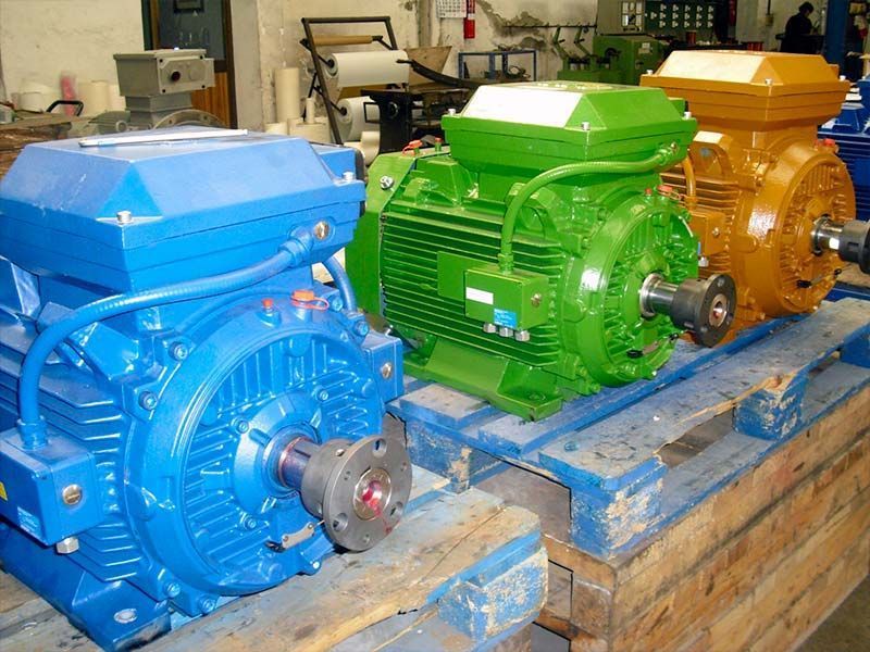 Tres motores eléctricos industriales de color azul, verde y amarillo, colocados uno al lado del otro sobre palés de madera en un almacén.