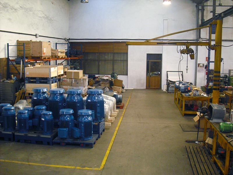 Interior de un almacén con filas de motores industriales azules, cajas de madera en estantes y un banco de trabajo con un polipasto.