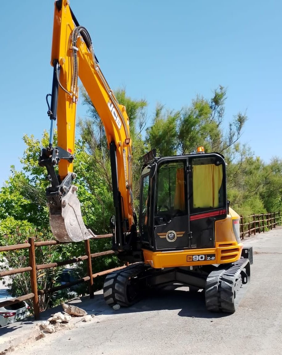 Excavadora amarilla en una carretera pavimentada al lado de una valla de madera y vegetación contra un cielo azul.