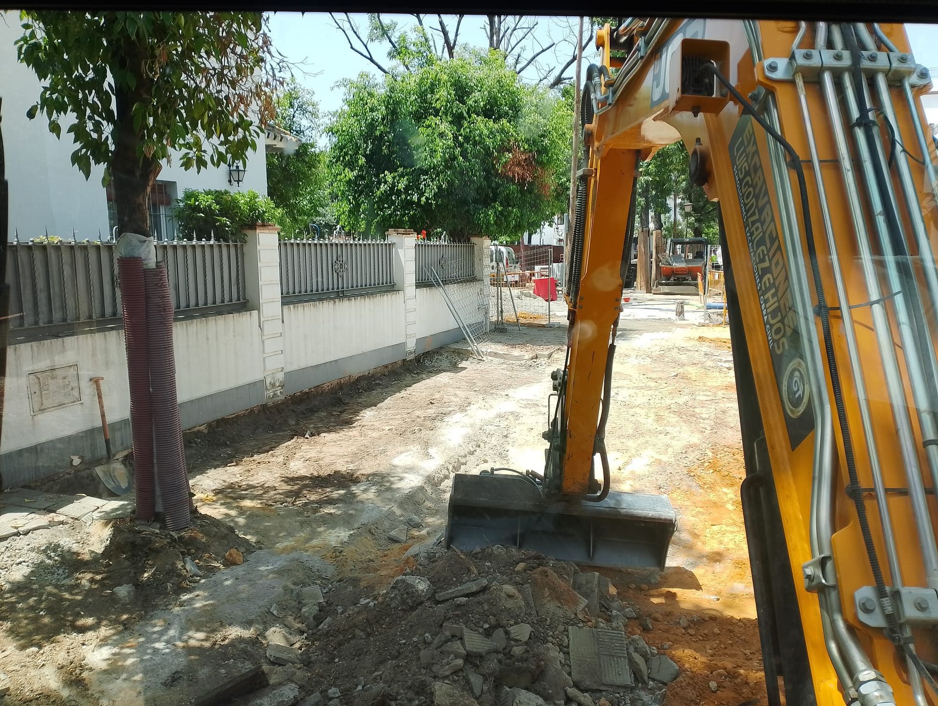 Excavadora cavando cerca de una valla y árboles en un día soleado.