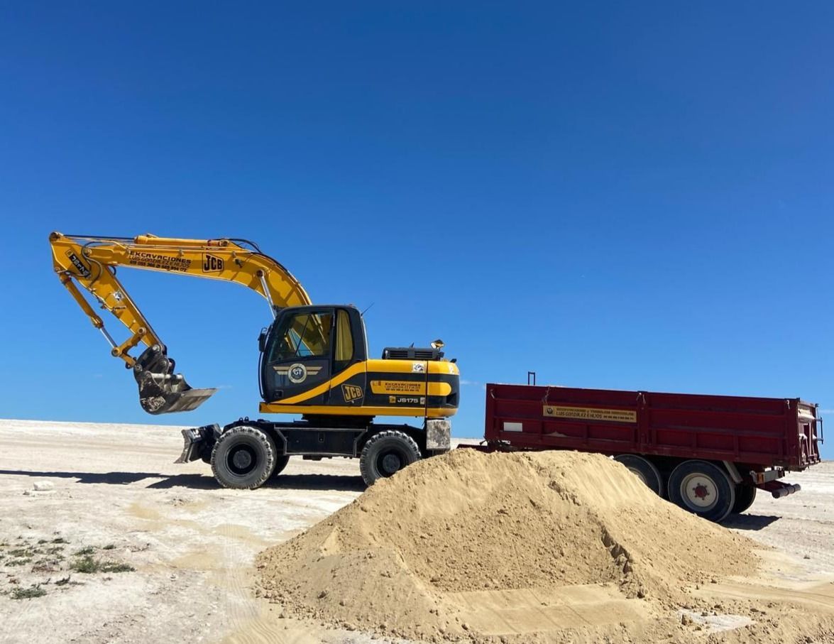 Excavadora amarilla cargando arena en un remolque rojo contra un cielo azul claro.