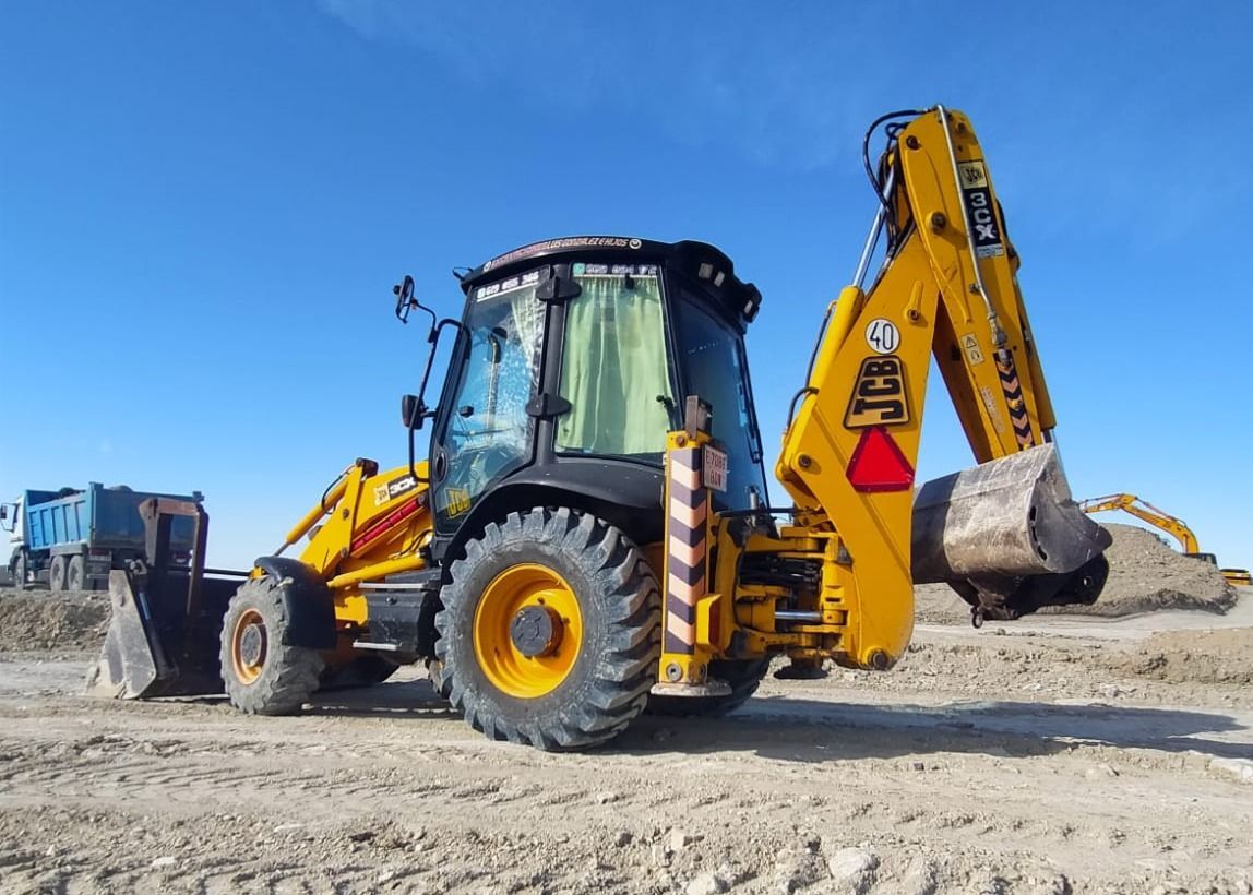 Retroexcavadora amarilla en una obra en construcción, bajo un cielo azul, con el cucharón hacia abajo.