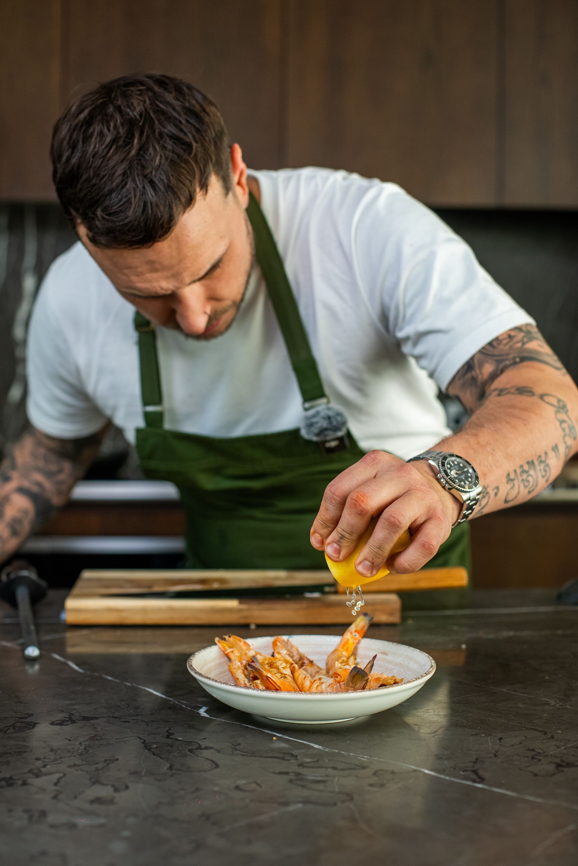 Chef in a white shirt and green apron squeezing lemon juice over shrimp in a white bowl.