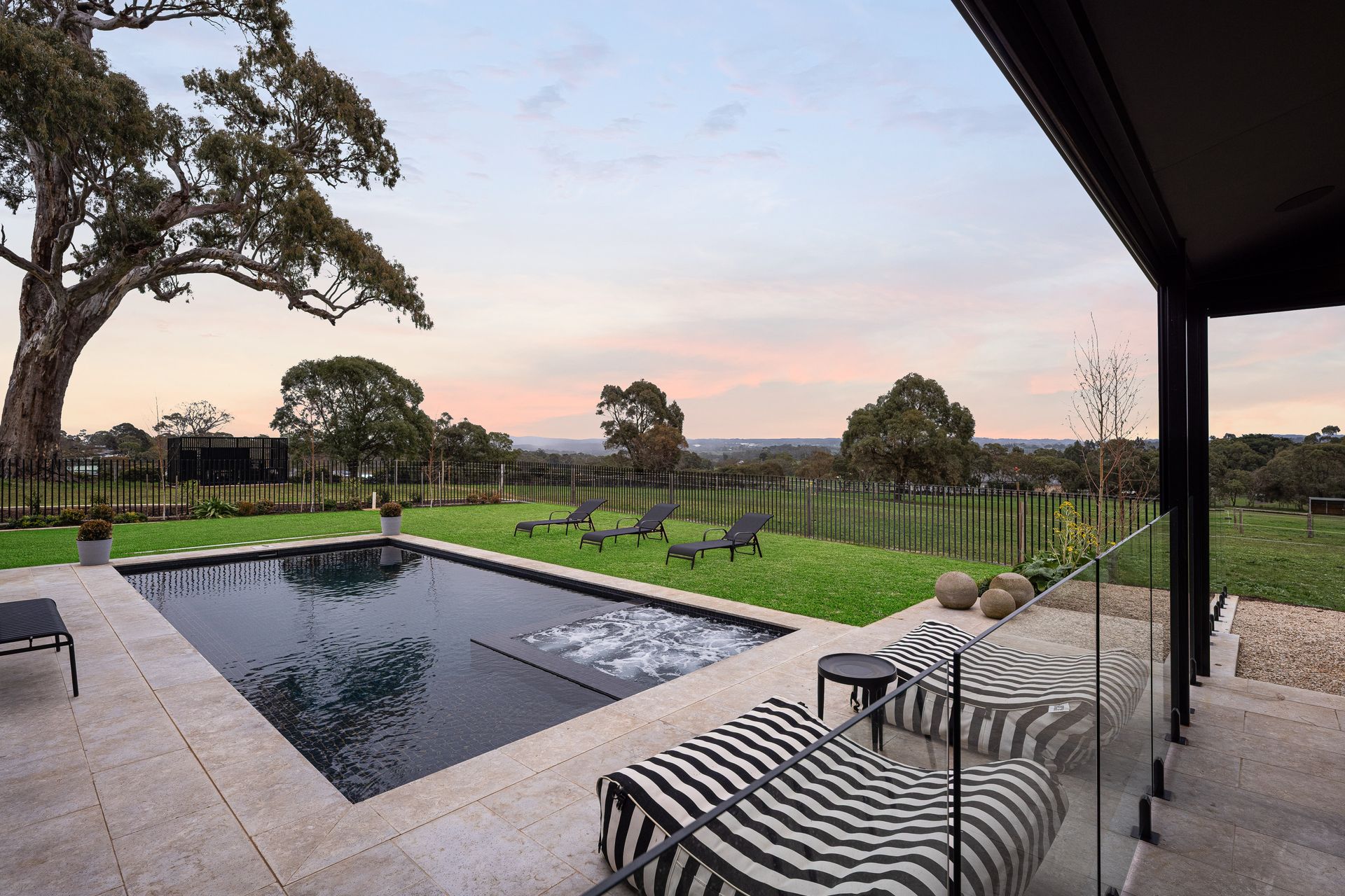 Swimming pool, lawn, and zebra-striped lounge chairs overlooking a rural landscape at dusk.