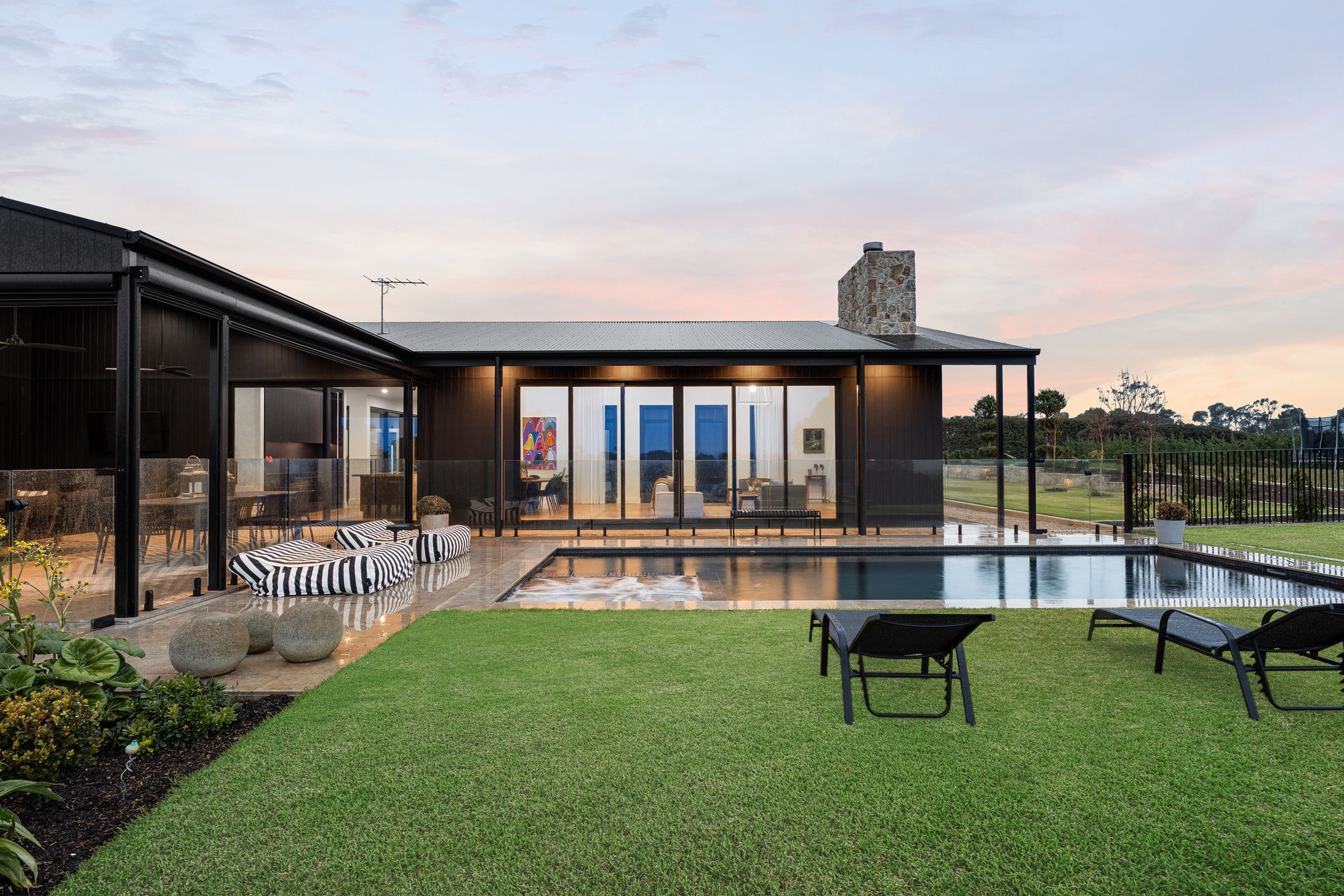 Modern house with pool, dark exterior, black lounge chairs on green lawn under dusk sky.