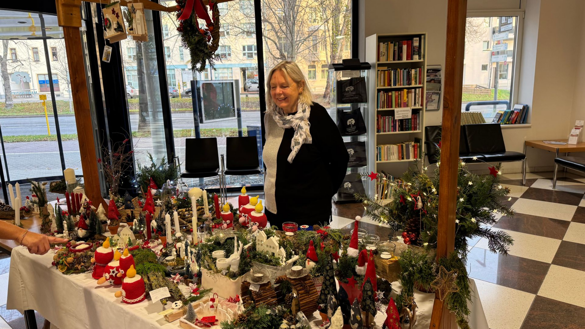 Frau an einem Marktstand mit Weihnachtsdekoration, lächelnd. Innenaufnahme mit Bücherregalen im Hintergrund.