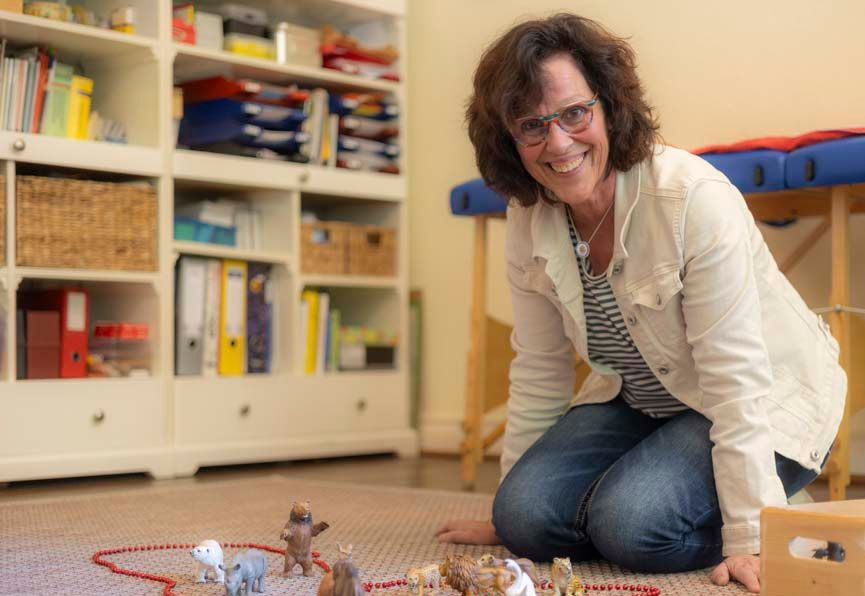 Eine Frau kniet lächelnd auf dem Boden, umgeben von Tierfiguren, in einem Raum mit Regalen und einer Massageliege.