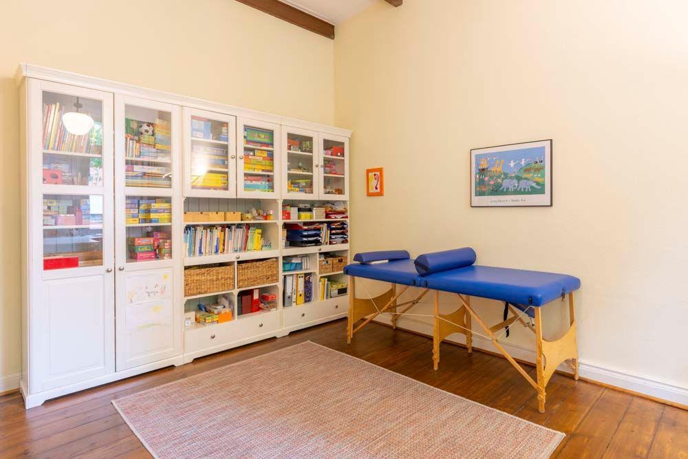 Zimmer mit Massageliege und weißem Schrank voller Utensilien. Beiger Teppich auf Holzboden.