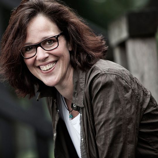 Frau mit Brille lächelt breit im Freien, trägt braune Jacke und weißes Hemd.