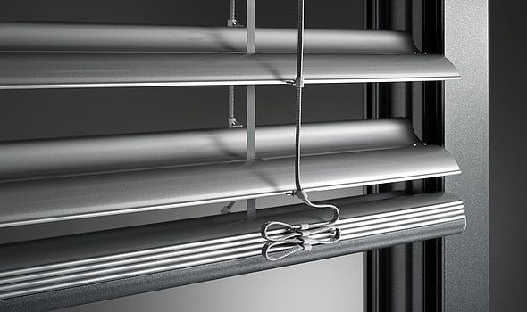 Nahaufnahme von silbernen Metalljalousien mit abgewinkelten Lamellen, die ein Fenster teilweise abdecken.